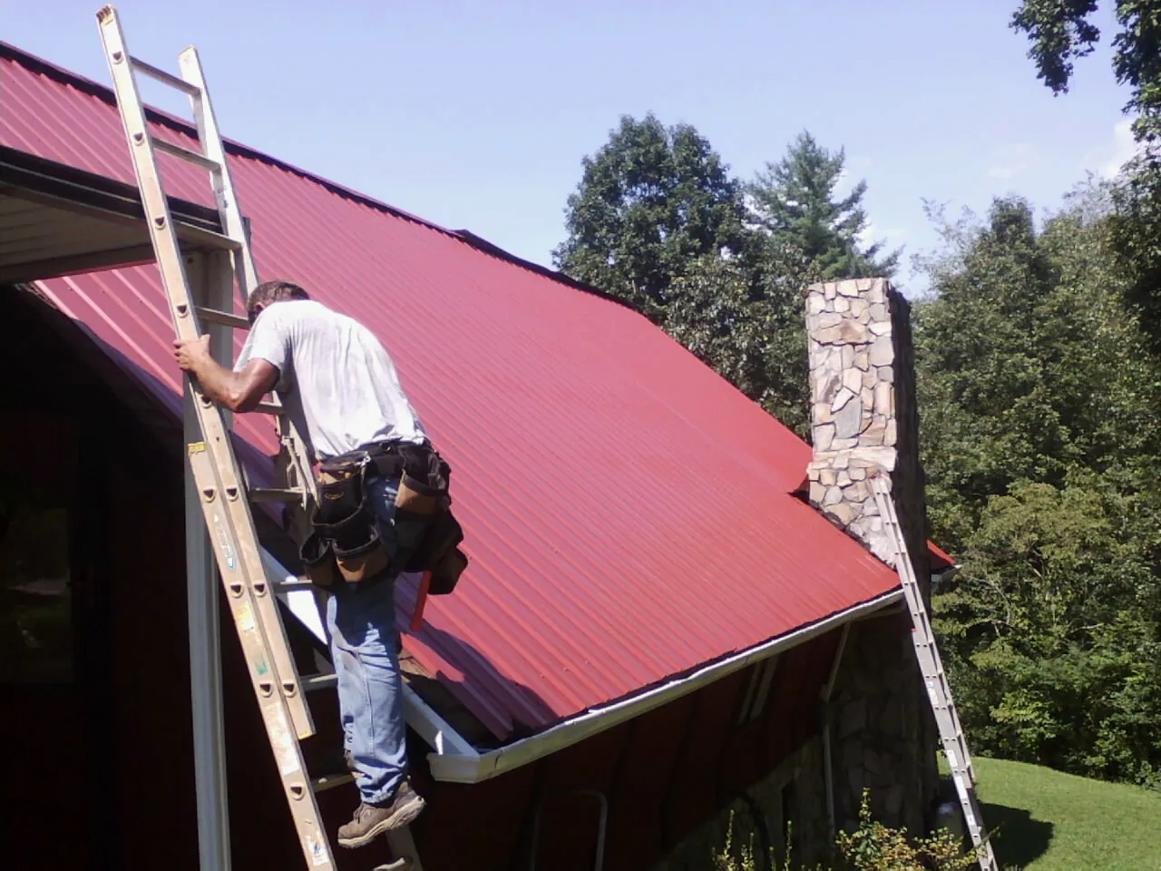 Roofer on a ladder beside a red metal roof and stone chimney — Soffit & Fascia Repair by Roofing Brothers in Coeur d'Alene
